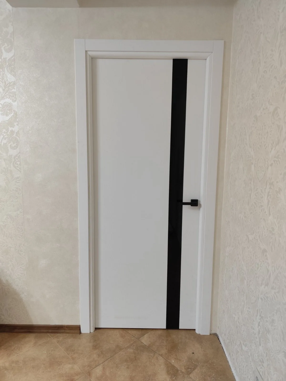 Ușă Plato Line 04 albă mată, cu o fâșie lată de sticlă neagră, între pereți cu tapet bej cu model