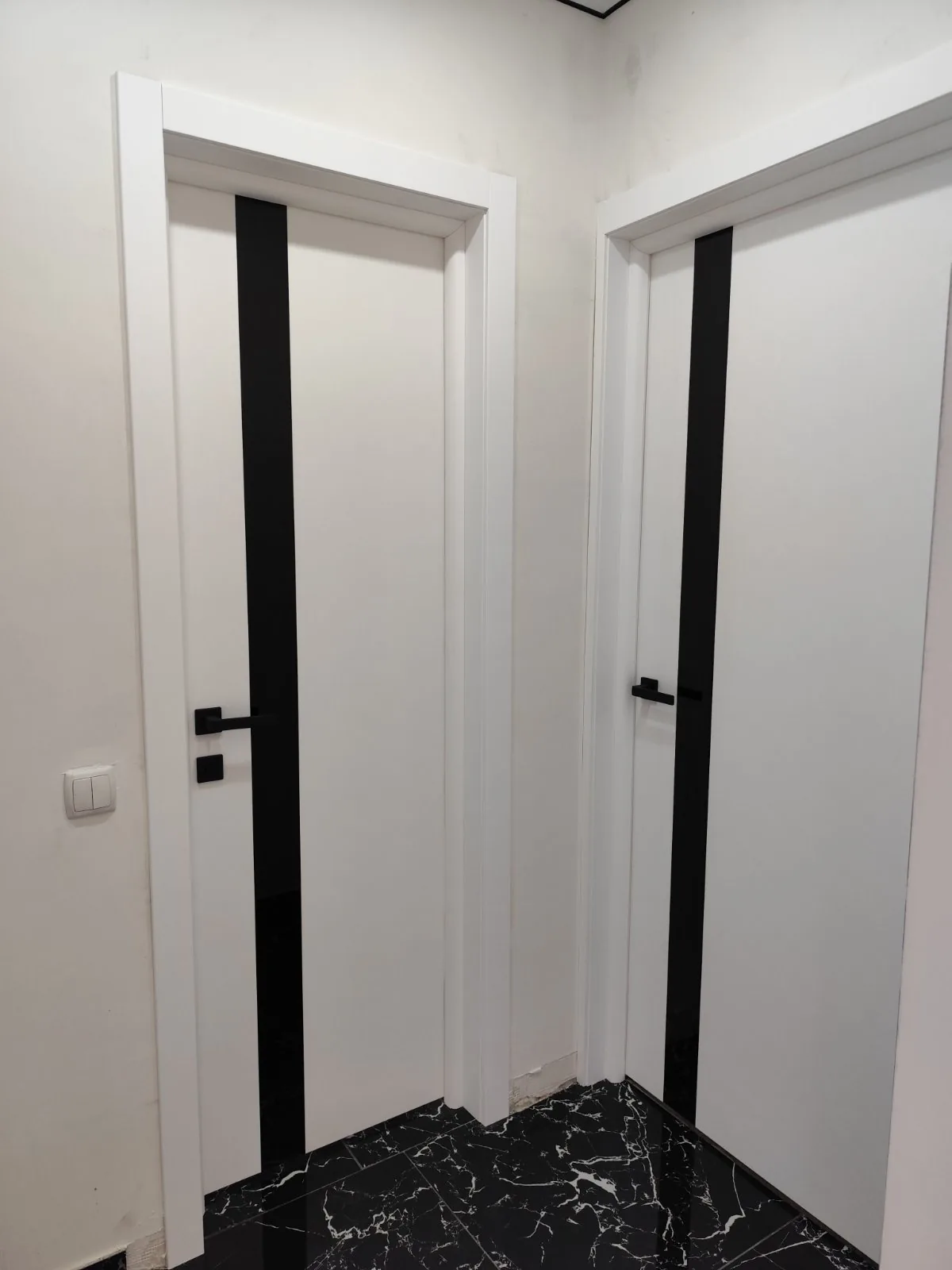 Două uși Plato Line 04 albe mate, cu sticlă neagră și mânere negre, pe o gresie neagră lucioasă