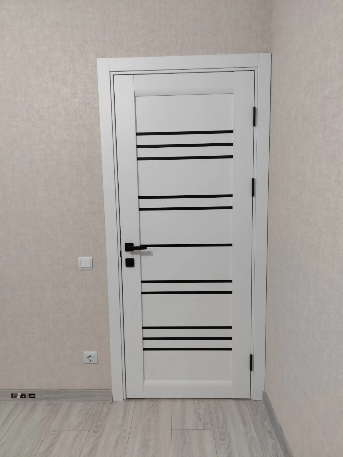 Ușă Versal albă texturată, cu grupuri de dungi de sticlă neagră și mâner negru, pe un perete cu tapet bej