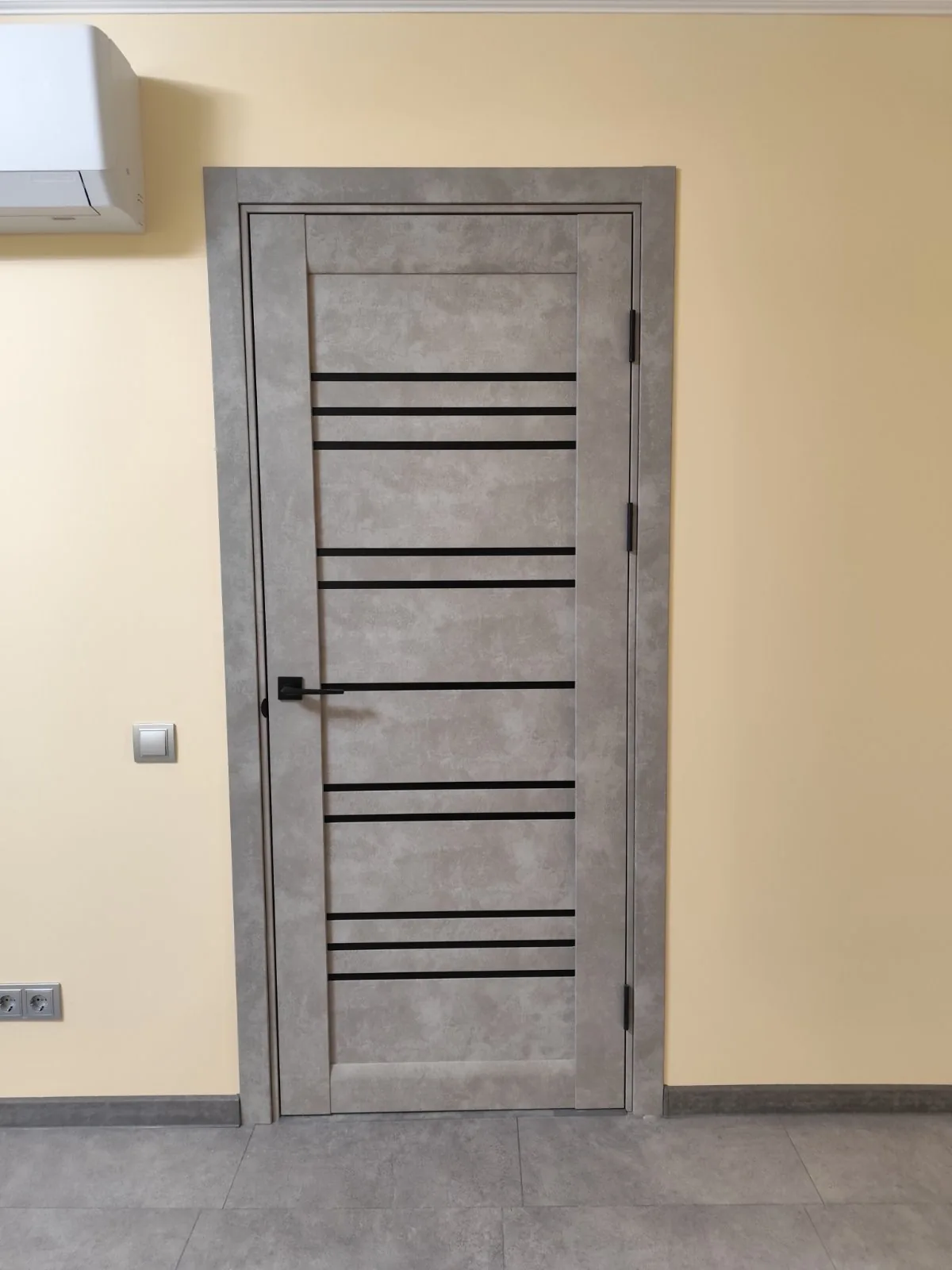 Ușă Versal gri beton, cu dungi de sticlă neagră și mâner negru, pe un perete galben pal